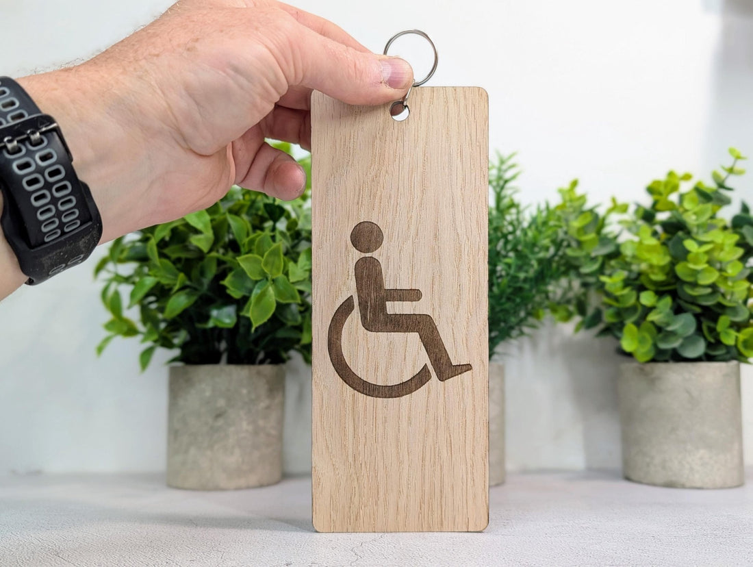 Hand holding a wooden keychain with a wheelchair symbol in front of plants