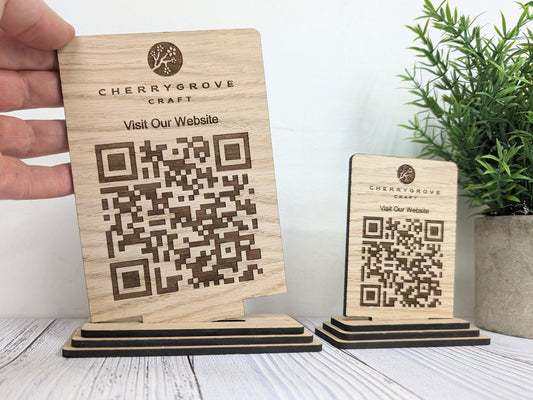 Large and standard freestanding oak QR code signs engraved with a logo and Visit Our Website, the large one held in a hand