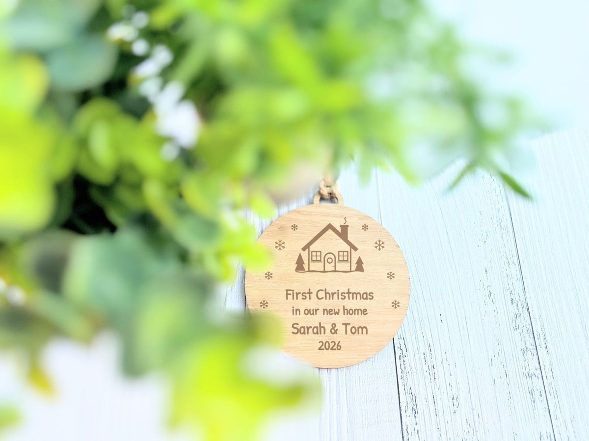 Personalised new home Christmas bauble in oak, engraved with names and year, shown with greenery