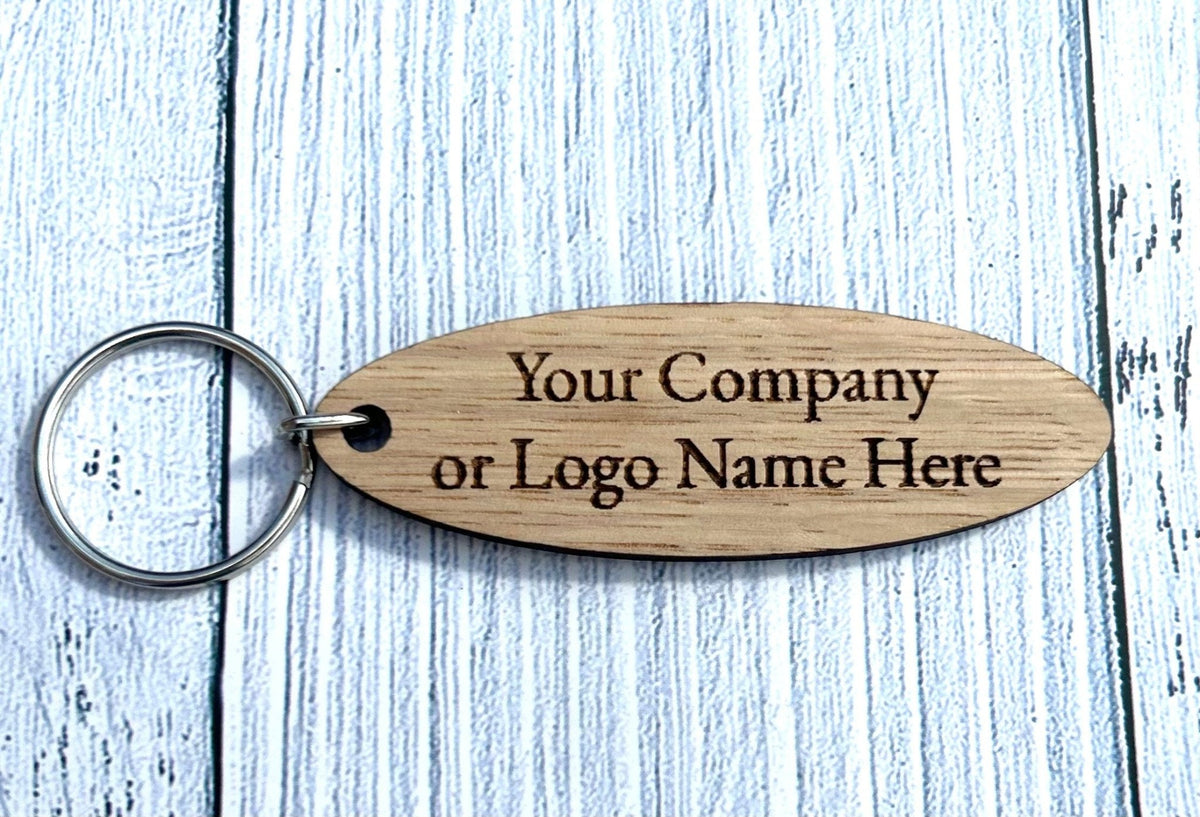 Laser-engraved oval oak keyring 75x25mm — warm blonde oak veneer MDF, recessed company name in crisp engraved lettering, silver split ring attachment, white background flat lay